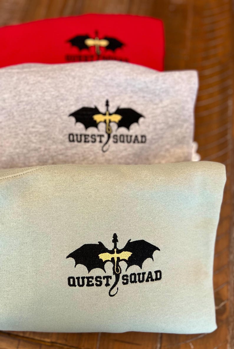 May include: Three sweatshirts stacked on top of each other. The top sweatshirt is red, the middle sweatshirt is gray, and the bottom sweatshirt is light green. Each sweatshirt has a black dragon with yellow wings and the text "QUEST SQUAD" embroidered on the front.