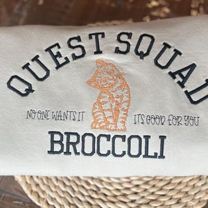 May include: A white sweatshirt with the text "QUEST SQUAD" in black embroidery. Below the text is a small orange and white cat illustration. The text "NO ONE WANTS IT" is on the left and "ITS GOOD FOR YOU" is on the right. The text "BROCCOLI" is below the cat illustration.