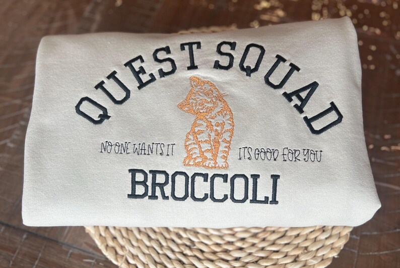 May include: A white sweatshirt with the text "QUEST SQUAD" in black embroidery. Below the text is a small orange and white cat illustration. The text "NO ONE WANTS IT" is on the left and "ITS GOOD FOR YOU" is on the right. The text "BROCCOLI" is below the cat illustration.
