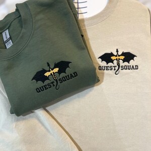 May include: Two sweatshirts, one in olive green and one in beige, both featuring an embroidered black dragon with gold accents and the text "QUEST SQUAD".