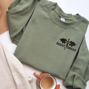 May include: A green sweatshirt with a black dragon graphic and the text "QUEST SQUAD" on the front.
