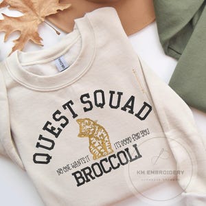 May include: A cream colored sweatshirt with black text that reads "Quest Squad" and "No one wants it, it's good for you, Broccoli". The text is surrounded by a black circle. A gold glitter cat is in the center of the circle.