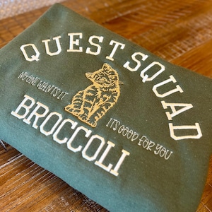 May include: Green sweatshirt with white embroidered text that reads "Quest Squad" and "No one wants it Broccoli it's good for you". A gold embroidered cat is sitting in the middle of the text.