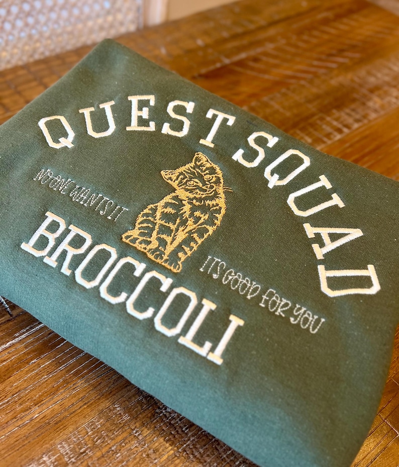 May include: Green sweatshirt with white embroidered text that reads "Quest Squad" and "No one wants it Broccoli it's good for you". A gold embroidered cat is sitting in the middle of the text.