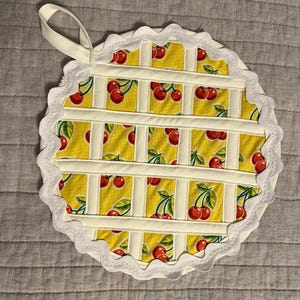 May include: Round, quilted pot holder with a yellow cherry print. The pot holder has a white scalloped edge and a loop for hanging. The cherry pattern is on a yellow background, with white vertical and horizontal strips creating a grid pattern.