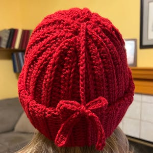 以下が含まれることがあります： 手作りの赤いかぎ針編みのビーニー帽、リブ編みデザイン。ビーニー帽の後ろには装飾的なリボンが付いています。この暖かい冬の帽子は寒い季節に最適で、どんな服装にも手作りの魅力を添えます。