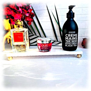 May include: A white marble tray with gold accents holds a clear glass bottle of perfume, a red jar of Olay Regenerist moisturizer, and a black bottle of Compagnie de Provence Creme Mains The Noir hand cream with black tea.
