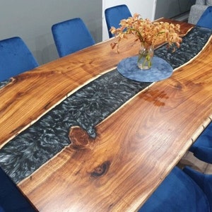 May include: A rectangular dining table featuring a natural wood grain surface with a live edge and a central inlay of black marbled epoxy resin. The table is surrounded by six blue velvet upholstered chairs and holds a glass vase with flowers.
