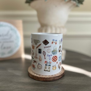 May include: White ceramic mug with a pattern of various illustrations, including a teacup, a book, a pie, a pair of boots, a bird, a rolling pin, a horseshoe, a mirror, a bow, a basket of flowers, a candle, a beehive, a watering can, a wheelbarrow, a spool of thread, a heart, a clock, and a bunch of flowers. The mug is sitting on a wooden coaster.