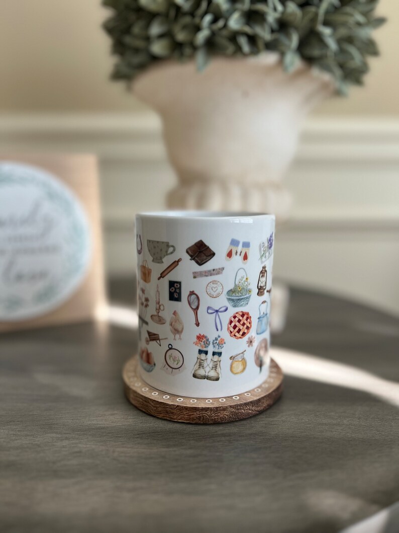 May include: White ceramic mug with a pattern of various illustrations, including a teacup, a book, a pie, a pair of boots, a bird, a rolling pin, a horseshoe, a mirror, a bow, a basket of flowers, a candle, a beehive, a watering can, a wheelbarrow, a spool of thread, a heart, a clock, and a bunch of flowers. The mug is sitting on a wooden coaster.