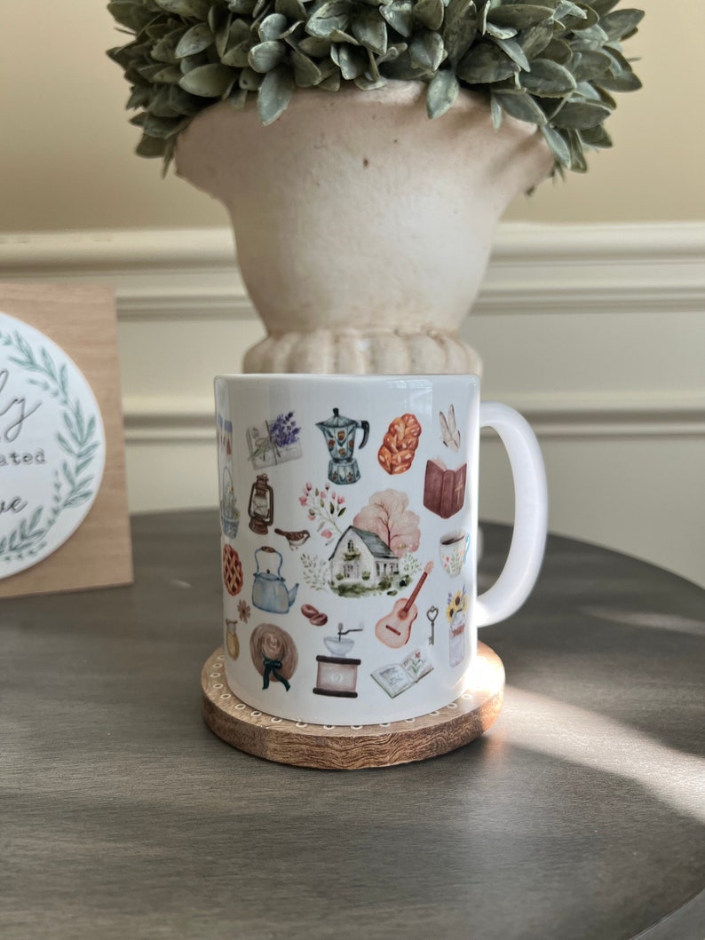 May include: White ceramic mug with a colorful illustration of various objects, including a house, a guitar, a teapot, a coffee grinder, and a book. The mug is sitting on a wooden coaster.
