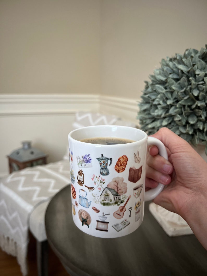 May include: A white ceramic mug with a handle, filled with coffee. The mug is decorated with a colorful pattern of various objects, including a house, a teapot, a guitar, and a bird.