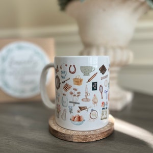 May include: White ceramic mug with a handle, featuring a pattern of various illustrations in shades of brown, blue, and green. The illustrations include a horseshoe, a teacup, a rolling pin, a basket, a wheelbarrow, a chicken, and other items. The mug is sitting on a wooden coaster.