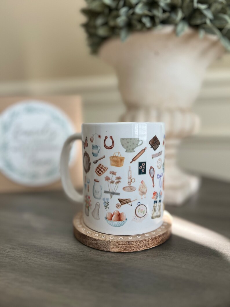 May include: White ceramic mug with a handle, featuring a pattern of various illustrations in shades of brown, blue, and green. The illustrations include a horseshoe, a teacup, a rolling pin, a basket, a wheelbarrow, a chicken, and other items. The mug is sitting on a wooden coaster.
