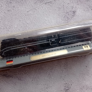 Può includere: Un locomotore di treno modello nero in una teca di plastica trasparente con un'etichetta dorata che recita "DRG Class 01-10". La teca include anche una piccola bandiera tedesca.