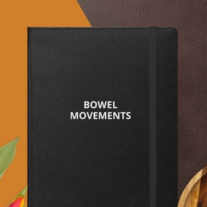 May include: A black leather-bound journal with the title "BOWEL MOVEMENTS" printed in white on the front cover. The journal is closed and lying on a brown surface.