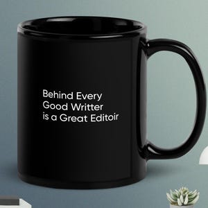 Peut inclure: Un mug en céramique noir avec une grande anse. Le mug présente le texte blanc : "Behind Every Good Writter is a Great Editoir". Le mug est posé sur une surface grise avec une petite lampe et une plante en pot.