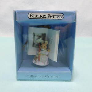 May include: A Beatrix Potter collectible ornament featuring a small mouse figurine wearing a light-colored dress and apron, standing in front of an open book. The ornament is encased in a clear plastic box with blue accents and text.