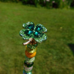 May include: A decorative topper featuring a four-leaf clover design adorned with dark green faceted stones and small light green crystals. The clover is mounted on a vertical rod decorated with a series of stacked, multi-colored glass beads.