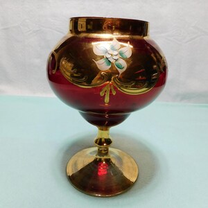 May include: A deep red glass vase with a wide, rounded body and a pedestal base. The upper portion features a metallic gold finish with decorative scrollwork and a hand-painted white flower with green accents centered on the front of the glass.