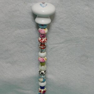 May include: A decorative pen featuring a white ceramic knob top with a small blue heart design. The shaft is adorned with a vertical arrangement of multi-colored beads, including floral patterns, solid blue, pink, and clear crystal-like accents.