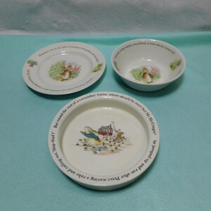 May include: A set of three white ceramic dishes featuring Peter Rabbit illustrations and printed text. The collection includes a small plate, a bowl, and a larger shallow dish, each decorated with colorful scenes of the rabbit in a garden setting.