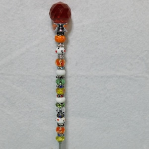 May include: A decorative wand with a red glass orb at the top and a variety of colorful beads, including orange, white, green, and yellow.