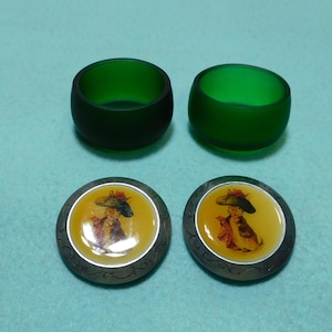 May include: Four decorative items: two green glass napkin rings and two round buttons with a yellow center featuring a vintage illustration of a person wearing a hat. The buttons have a silver-colored border.