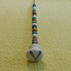 May include: A long metal needle topped with a white ceramic knob featuring a graphic of two crossed scissors. The needle shaft is decorated with a vertical row of multi-colored beads in shades of blue, orange, white, and silver, arranged in a pattern.