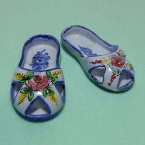 May include: Pair of blue and white ceramic slipper-shaped trinket dishes with floral designs. The dishes are painted with pink, yellow, and blue flowers.