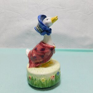 May include: A ceramic duck figurine wearing a blue bonnet and a red shawl with dark spots. The duck stands on a cylindrical base decorated with green grass and small colorful flowers, featuring a white beak and detailed painted features.