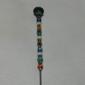 May include: A long, silver metal rod with a teal ceramic top and colorful beads. The beads are in various colors, including blue, green, orange, white, and yellow.