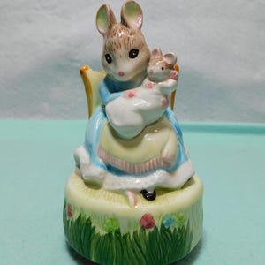 May include: A ceramic figurine of an adult mouse sitting in a chair, holding a smaller baby mouse wrapped in a white blanket. The adult mouse wears a blue dress with a yellow apron. The base is painted with green grass and small flowers.
