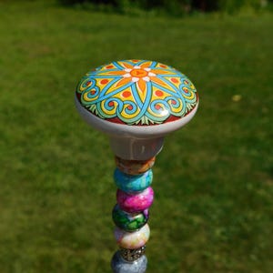 May include: A decorative garden stake featuring a round ceramic knob with a colorful orange, yellow, and blue floral pattern. The stem is adorned with a vertical stack of multi-colored, marbled glass beads in shades of pink, green, blue, and white.