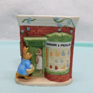 May include: A ceramic figurine of a shop front with red brick walls and a green door. A rabbit in a blue jacket stands outside. The shop window displays the text GINGER & PICKLES with various colored bottles inside. Blue flowers climb the wall.