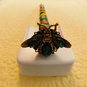 May include: A decorative pen featuring a gold-toned bee topper with dark green oval-shaped stones on its wings and body. The pen barrel is adorned with a series of colorful, patterned beads in shades of green, yellow, orange, and red.