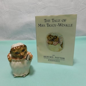 May include: A ceramic hedgehog figurine wearing a white cap and a plaid shirt, standing next to a book titled The Tale of Mrs. Tiggy-Winkle by Beatrix Potter, published by F. Warne & Co. The book cover features an illustration of a hedgehog.