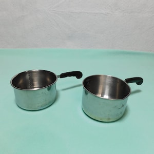 May include: Two silver measuring cups with black handles. The cups are marked with measurements in ounces and milliliters.