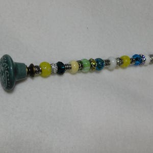 May include: A teal ceramic knob with a silver metal connector attached to a beaded string. The string is made of various colored beads, including yellow, green, blue, and white.