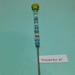 May include: A decorative threaded rod with a lime green top and a series of blue, white, and silver beads. The beads are of various sizes and designs. The label reads "Threaded Rod #7".
