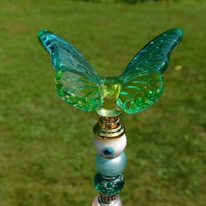 May include: A decorative topper featuring a translucent green glass butterfly with intricate wing patterns, mounted on a gold-toned metal base. Below the butterfly is a vertical stack of assorted beads, including white, blue, and pink glass spheres.