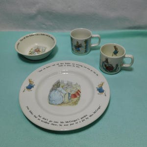 May include: A ceramic dinnerware set featuring Peter Rabbit illustrations and text. The set includes a white plate, a bowl, and two mugs. The plate features a central scene of rabbits and text around the rim. The bowl and mugs also display character graphics.
