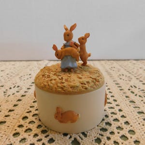 May include: A round ceramic trinket box with a cream-colored base featuring a small brown rabbit in relief. The lid has a textured tan surface topped with a figurine of a larger rabbit in a blue dress holding a smaller rabbit, with another small rabbit standing beside them.