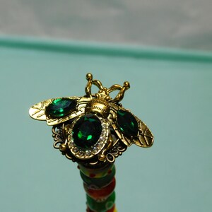May include: A gold-toned bee-shaped decorative topper featuring two marquise-cut emerald green stones on the wings, a central oval-cut emerald green stone surrounded by a halo of clear crystals, and a textured metal body with antennae.