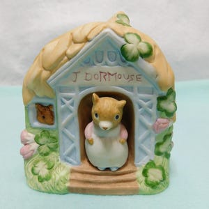 May include: A ceramic figurine of a house with a thatched roof and light blue walls, featuring the text J DORMOUSE above the doorway. A small brown dormouse figure stands in the entrance, while another dormouse face appears in a side window.
