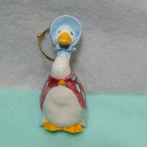May include: A ceramic goose ornament featuring a white body, a yellow beak, and yellow feet. The goose wears a light blue bonnet and a red cape with light blue polka dots, fastened with a yellow button. A gold hanging cord is attached to the top.