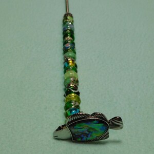 May include: A decorative metal pin featuring a vertical row of multi-colored green, yellow, and blue glass beads interspersed with small silver-toned spacers, terminating in a silver-toned fish charm with an iridescent abalone shell inlay.