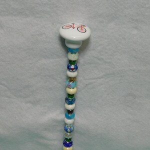 May include: A decorative garden stake featuring a white ceramic knob top with a red bicycle graphic. The stem is adorned with a vertical arrangement of multi-colored glass and metallic beads in shades of blue, green, white, and yellow, creating a vibrant, textured column design.