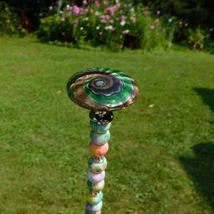 May include: A decorative garden stake featuring a large, flat, circular glass topper with a swirling green, black, and copper-toned pattern. Below the topper, a vertical rod is adorned with a series of small, multi-colored, patterned glass beads.
