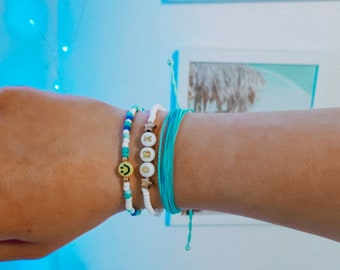 May include: A person's wrist wearing four bracelets. The bracelets are made of white beads, blue beads, and a turquoise string. One bracelet has a smiley face charm and another has the letters "XO" in gold.